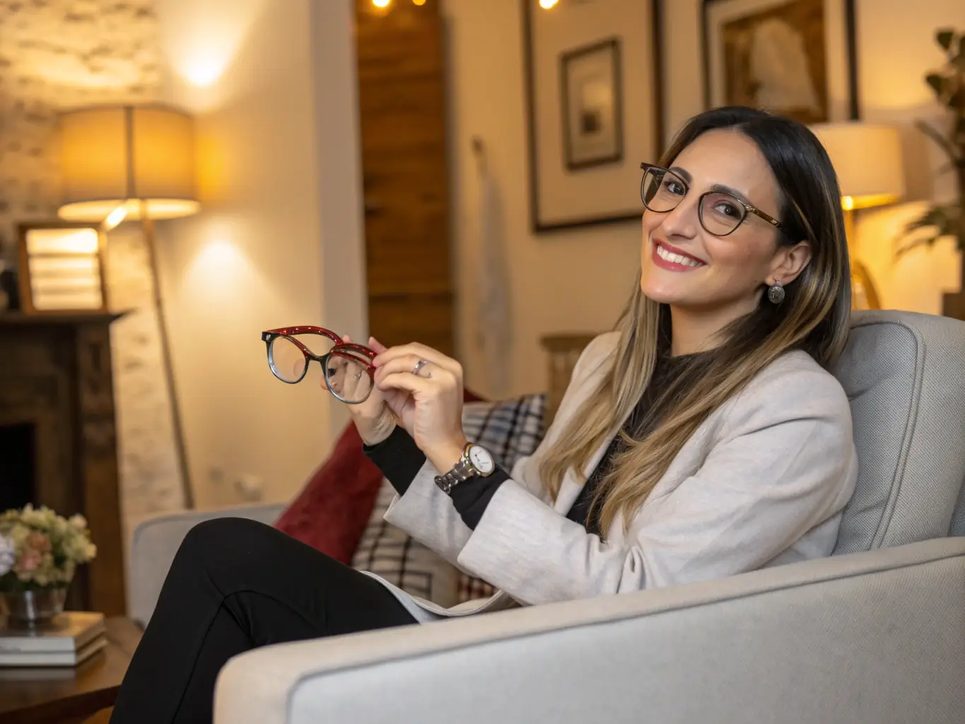 A friendly optician technician, Lorena, is smiling and holding a frame selection kit in a modern living room setting, ready to assist a client with choosing the perfect frame.